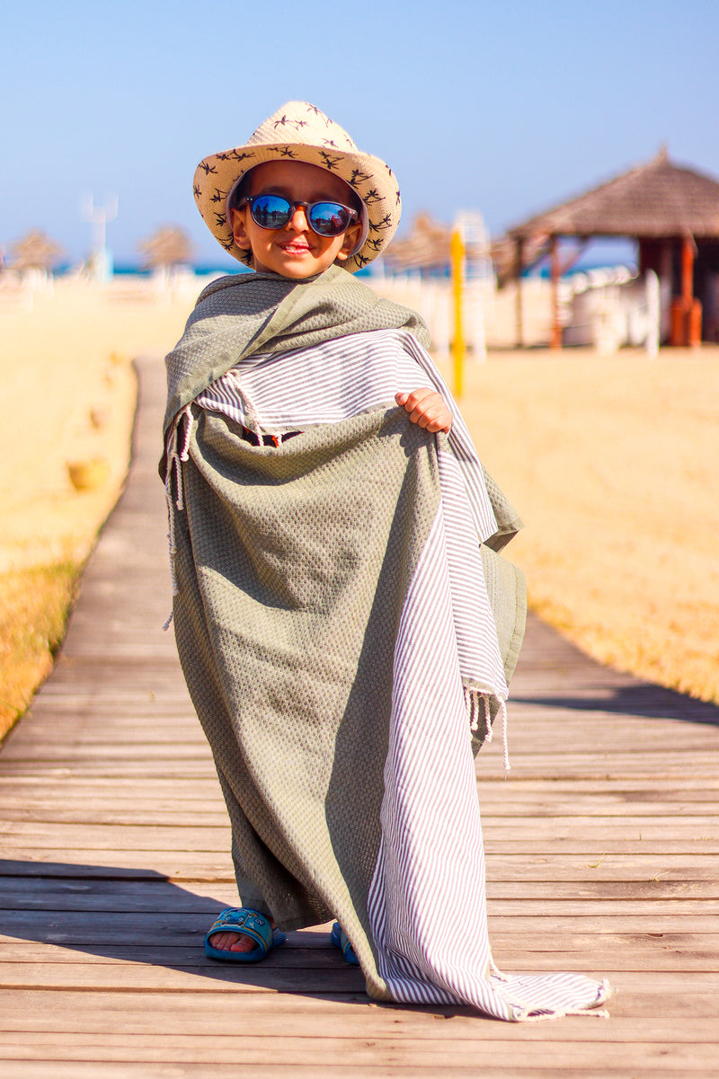 kind mit eingewickeltem Strandtuch Hamamtuch am Strand Strohhut und Sonnenbrille tragend