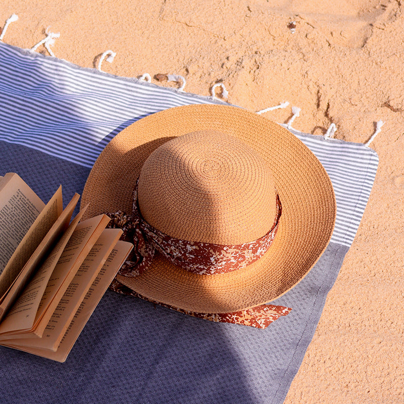 eine graue Hamamtuch Fouta am Strand mit einem Buch und einem Hut