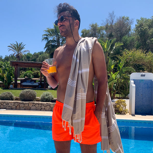 ein Mann am Pool mit einem Glas Saft in der Hand und einem beigen Strandtuch auf den Schultern