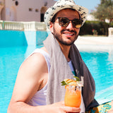 Mann mit Sonnenbrille und Hut trägt hellgraues Strandtuch auf den Schultern am Pool