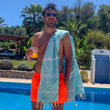 ein Mann am Pool mit einem Glas Saft in der Hand und einem aquablauen Hamamtuch auf den Schultern