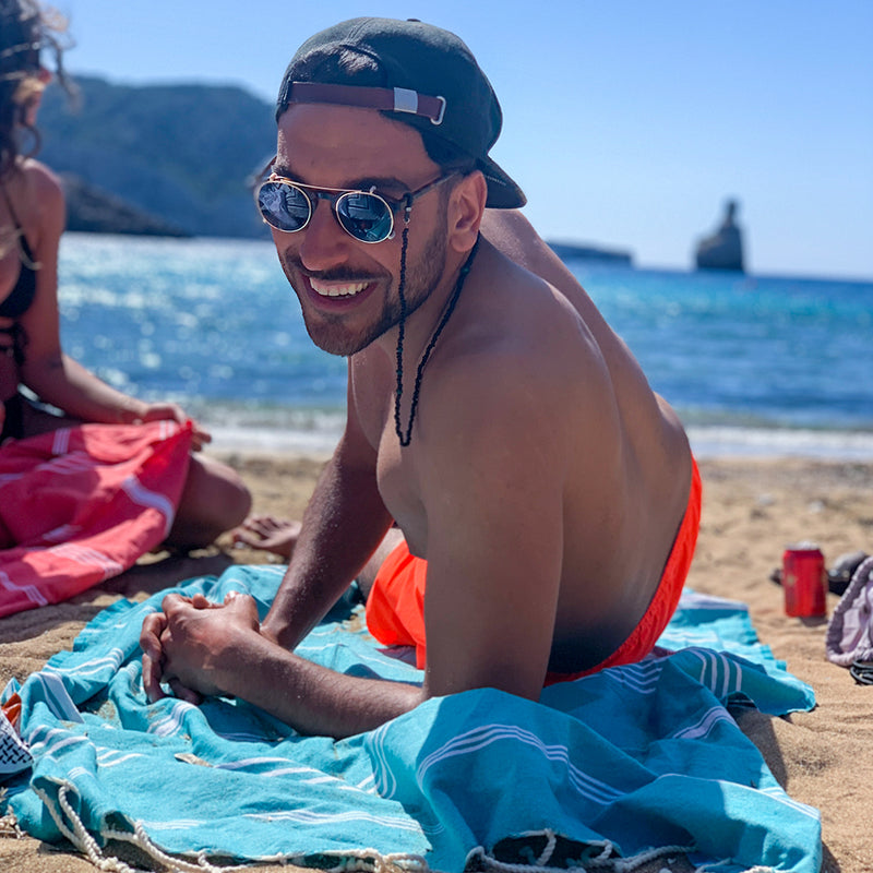 Mann mit Sonnenbrille auf einer hellblauen Hamamtuch am Strand liegend