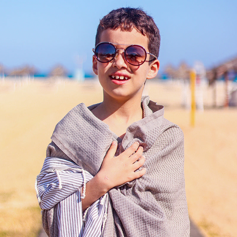 ein Kind mit Sonnenbrille und dunkelgrauem Strandtuch fouta auf den Schultern am Strand