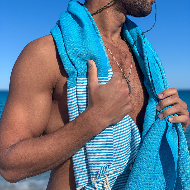 ein Mann am Strand der eine blaue Saunatuch Fouta auf den Schultern trägt