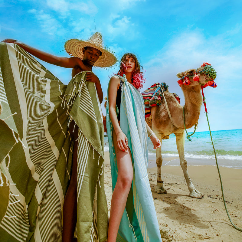 zwei Mädchen am Strand mit olivgrünem Strandtuch und aquablauer Fouta stehen neben einem Kamel