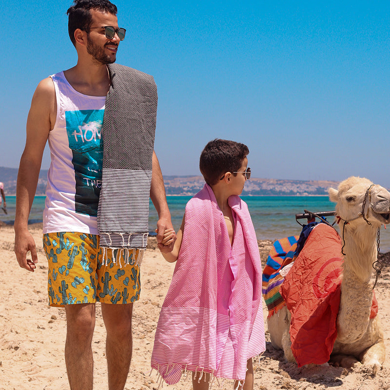 ein Vater in dunkelgrauer Fouta und der Sohn in rosa Saunatuch in der Nähe des Kamels am Strand