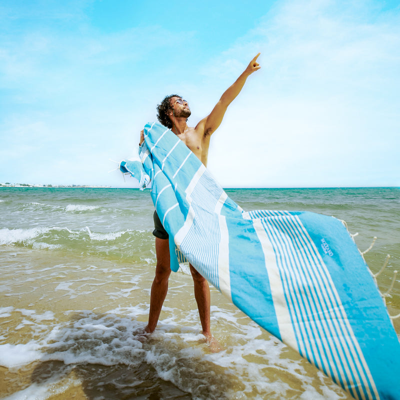 ein Mann am Strand der ein aquablaues Strandtuch Fouta hält