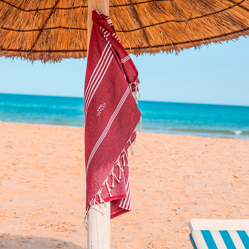 ein dunkelrotes Strandtuch fouta das unter einem Sonnenschirm am Strand hängt