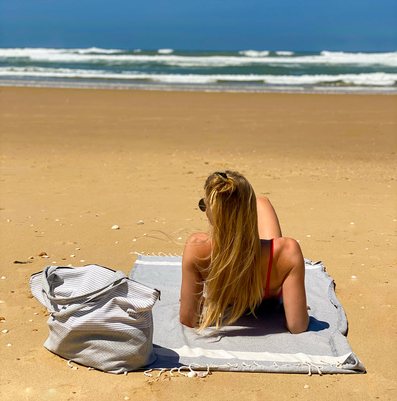 Mädchen mit Strandtasche und hellgrauem Strandtuch fouta am Strand liegend
