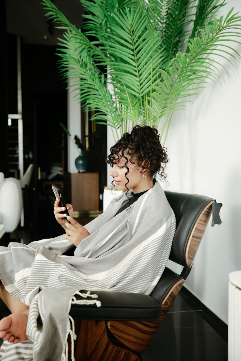ein Mädchen auf einem Liegestuhl mit pastellgrauer Decke auf den Schultern das mit seinem Handy spielt